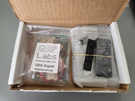 A small open shipping box containing several bags of electronic parts. One bag proclaims that it is a 'QDX Digital transceiver' from QRP Labs.