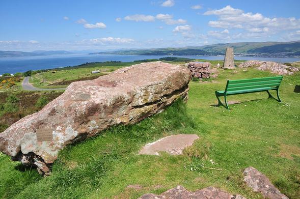 The highest point on the island of Great Cumbrae. The image shows a view taking in the distant Firth of Clyde with land visible extending across the upper part of the frame. In the foreground on the right is a trig point and a green bench, while on the left is a large whale-shaped rock in pinkish stone. The scene is in sunlight.