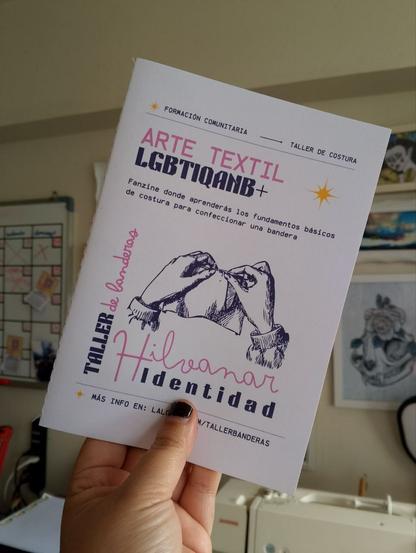 Una mano sostiene un fanzine titulado “Arte Textil LGBTIQANB+” con ilustraciones de manos cosiendo. El subtítulo dice “Fanzine donde aprenderás los fundamentos básicos de costura para confeccionar una bandera”. También se lee “Taller de banderas – Hilvanar Identidad”.