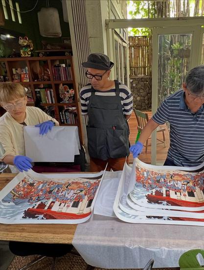photo of a group of 3 people standing over a table where there are copies of the art print mentioned in the previous description