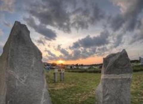 quasi-rural landscape with parked cars framed by two large stones in foreground, three informational markers visible in middle distance, sun on the horizon in the distance with a dramatically partially cloudy sky