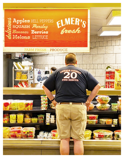 AI-assisted alt text:

A man in a DEKALB COUNTY FIRE RESCUE 20 shirt stands facing a refrigerated display of fresh fruit cups in a grocery store, hands on waist. Above him is an orange sign listing fruits and vegetables and reading ELMERS fresh SINCE 1928.