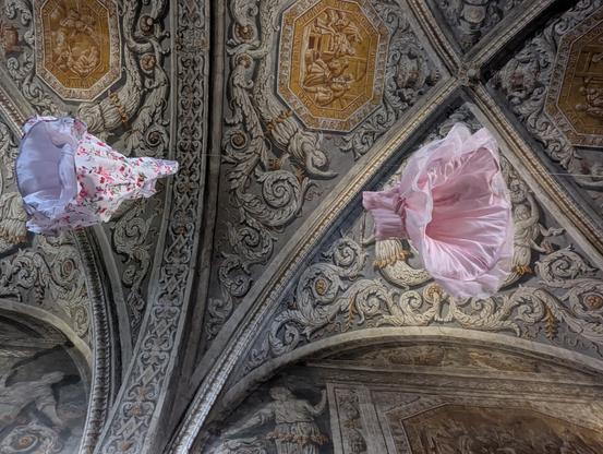 Deux petites robes d'enfants dans les tons rose pastel suspendues au plafond richement orné d'une chapelle. On les voit du dessous.