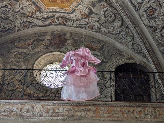 Robe d'enfant rose pastel et à froufrous suspendue au plafond d'une chapelle. À l'arrière, une balustrade en fer forgé, une porte et une fenêtre circulaire.
