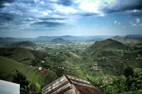 A panoramic view of the lush, rolling hills of Uganda under a dramatic, cloudy sky. The foreground shows the rusty, corrugated iron roof of a building and some green foliage. In the midground, the hills are terraced and dotted with small homes and agricultural plots, leading to a valley with what appears to be a village. Distant mountains fade into the haze on the horizon.