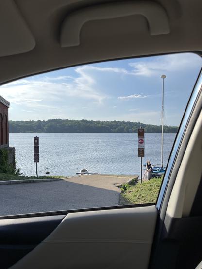 A view of Mystic Lake seen through the car window. In the foreground is a cement boat ramp curving down into the water. At the bottom right of the ramp a person is sitting on the gunnel of a catamaran-style sailboat. The surface of the water is rippled by a steady breeze. In the distance a row of trees at the far shoreline creates a horizon. The sky above is laced with swaths of cloud.