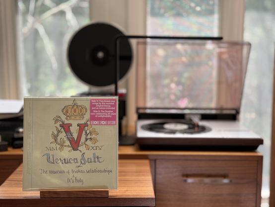 Veruca Salt - V (2014) LP cover.

A drawing of a V with a crown over it, surrounded by vines. The date, in roman numerals, is across the bottom of the V, left and right. Below, "Veruca Salt" and then the two tracks on the front - "The Museum of Broken Relationships" and "It's Holy".

The black 10" single plays on a vintage, restored Bang & Olufsen Beogram 4000 linear-tracking turntable to the right.