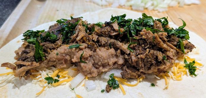 Burrito filling on top of a flour tortilla consisting of shredded cheese, white onion, refried beans, beef barbacoa and cilantro.