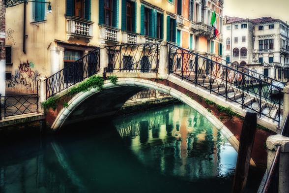 Eine kleine Brücke mit Treppen über einem Runden Bogen und mit einem dünnen schwarzen Geländer. Unter der Brücke fließt tief grünes Wasser in einem Kanal. Die Brücke ist teilweise grün bewachsen und führt links zu einem beigen historischen Gebäude mit kleinen Balkonen und grünen Fensterläden. Das Bild strahlt eine märchenhafte Atmosphäre aus.