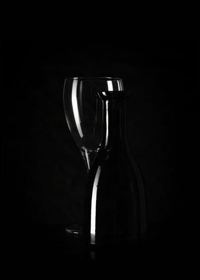 A wine glass and a bottle are set against a dark background. The glass is clear and slightly reflects light, positioned partially behind the black bottle, which has a matte finish. The composition creates subtle highlights and minimal shadow, emphasizing the contours of both objects.