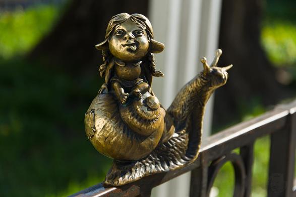The image captures a charming palm-size bronze sculpture of a young girl with curly hair, sitting on the back of a big snail. The snail's spiral shell provides a comfortable seat for the child showing her tongue as if having just accomplished something mischievous. The girls hair is styled in two braids, and her large drooping a bit pointy ears are clearly visible. The snail appears to be moving along what looks like a fence or railing made from dark metal with vertical bars.

The sculpture exhibits signs of age and weathering, which lend it an antique charm. Its patina indicates that the bronze has oxidized over time, giving it a rich coppery hue.

The background is softly focused but reveals hints of greenery which could suggest an outdoor setting like a garden or park where such sculptures are often found to enhance aesthetics while telling stories through art.