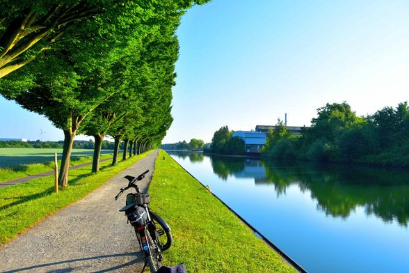 A bicycle rests on a gravel path beside a calm canal, lined with green trees and grass under a clear blue sky.
Ein Fahrrad steht auf einem Kiesweg neben einem ruhigen Kanal, gesaeumt von gruenen Baeumen und Gras unter einem klaren blauen Himmel.