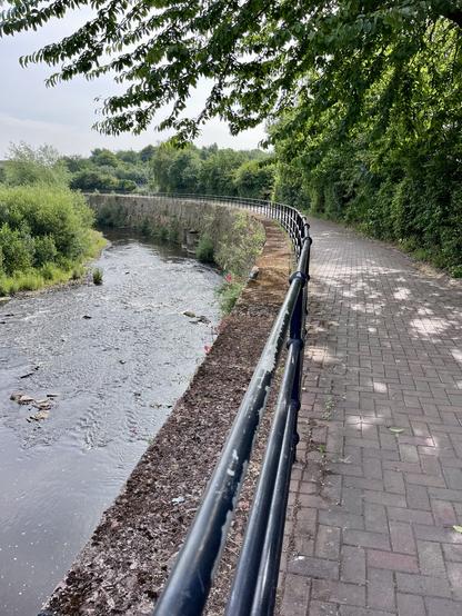 Paved footpath with a railing and retaining wall separating it from the river, flowing away from the camera. Both sides lined with green vegetation and the sky is a light cloudy blue