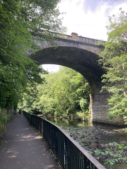A massive stone/brick bridge spanning a river, surrounded by trees and with a small person for scale