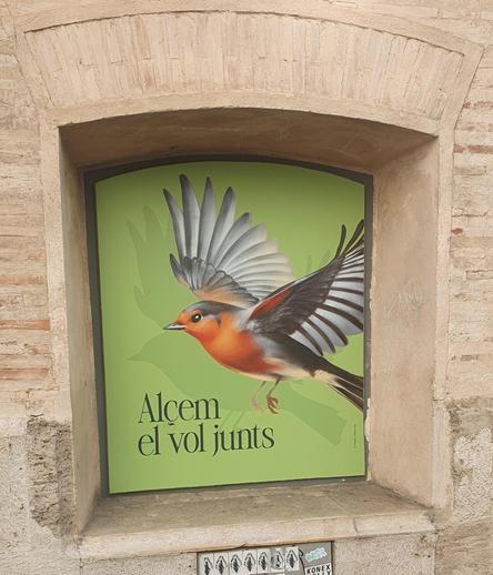 Cartell en la façana del Centre del Carme amb la imatge d'un ocell taronja sobre fons verd i el text "Alçem [sic] el vol junts"