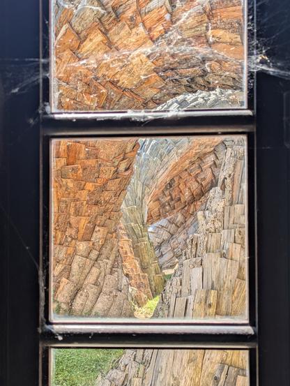 À travers les vitres carrées d'une  fenêtre, on voit une œuvre d'art en extérieur qui ressemble à de grosses racines couvertes d'une sorte d'écorce écailleuse en bois.