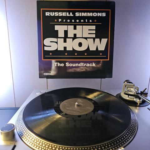 A vinyl record sits on a turntable. Behind the turntable, a vinyl album outer sleeve is displayed. The front cover shows the text "Russell Simmons Presents The Show The Soundtrack".