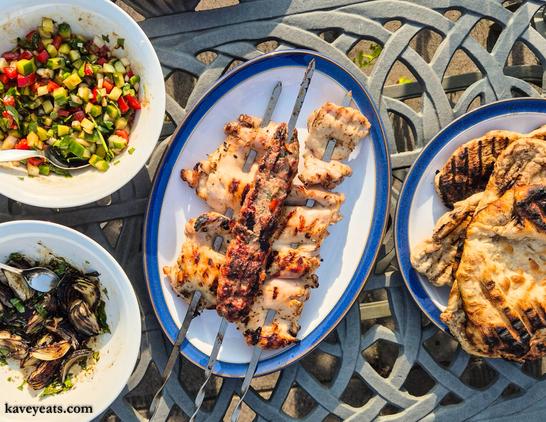 Photo of outdoor dining table with home booked recipes from the cookbook: marinated chicken thighs, lamb beyti, charred onions, shepherd's salad and flatbreads