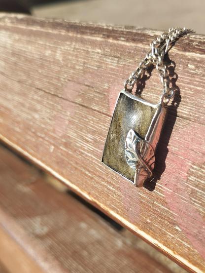 a silver pendant with a silver leaf with brown square gemstone. it's placed on blown wood