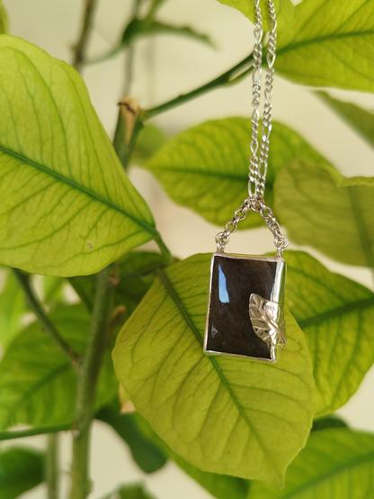 a silver pendant with a silver leaf with brown square gemstone. it's hanged from a silver chain. in the background green leaves