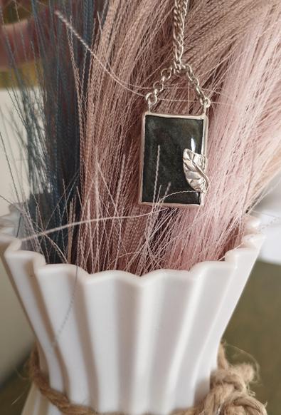 a silver pendant with a silver leaf with brown square gemstone. on the background a white base with grey plants