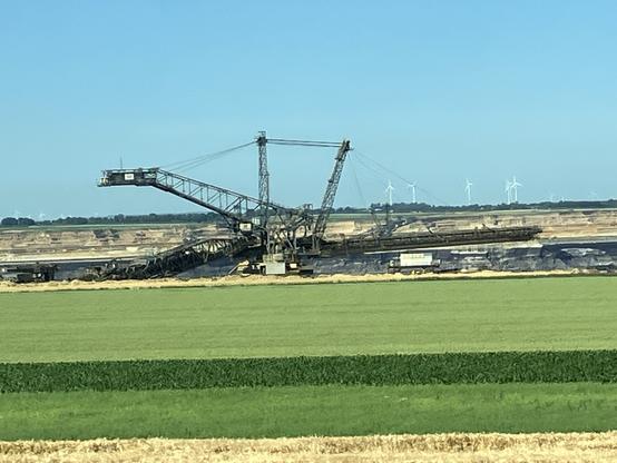 Im Vordergrund ein Feld. Zentral im Bild ein Braunkohlenbagger. dahinter die steilen, gelbbraunen kahlen Böschungen des Tagebaus. Am Horizont ein Dutzend Windräder.