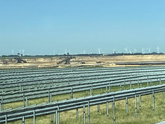 Im Hintergrund Braunkohletagebau mit Baggern. Am Horizont Windräder. Vorne viele Reihen von Alugestellen für Photovoltaik, noch ohne Module.