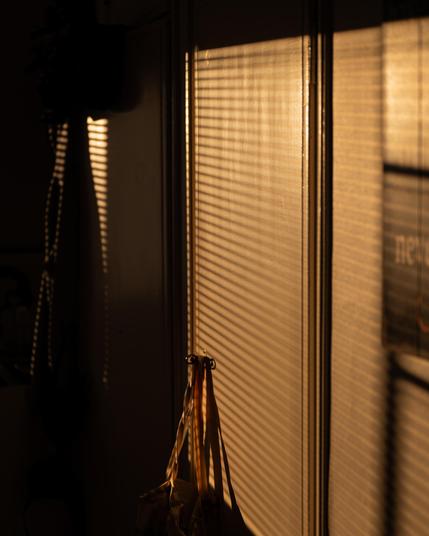 Vertical window blinds casting striped shadows on a wall, illuminated by warm sunlight. A bag hangs from a hook in the lower center, partially obscured by shadows. The lighting creates a contrast between the bright wall and dark areas.