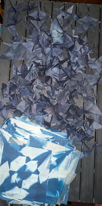 A table, dark grey and slatted, seen from above, with a big pile of dark blue-gray origami cranes on top. About 200 of them.
At the bottom is a pile of images on white paper which are blue geometric shapes. Each is what the origami cranes will look like when open and developed. There are about 400 of them, though only the top one is clearly visible