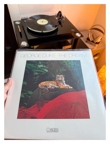 Auf einem schwarzen Plattenspieler läuft eine schwarze Schallplatte. Davor halte ich das Cover ins Bild. Es ist weiß, mittig ist ein gemaltes Bild eines Tigers, der auf einem Stein liegt, dahinter grüner Dschungel.
