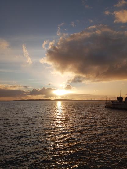 Una puesta de sol dorada sobre un cuerpo de agua tranquilo. El sol se encuentra en el horizonte, detrás de una línea de tierra oscura y plana, y sus rayos se reflejan en el agua, creando un brillo dorado. Las nubes en el cielo son principalmente de tonos dorados y anaranjados, con algunas áreas más oscuras. En la parte inferior derecha de la imagen, hay una parte de un paseo marítimo con farolas. La escena es pacífica y serena.