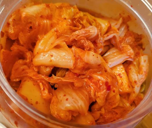 High-angle, close-up shot focuses sharply on the content of a clear plastic container filled with kimchi. The dominant colors are the reddish-orange from the sauce coating the light yellow and pale green of the kimchi cabbage. The composition is filled entirely with the kimchi and the container's rim, eliminating background distractions.