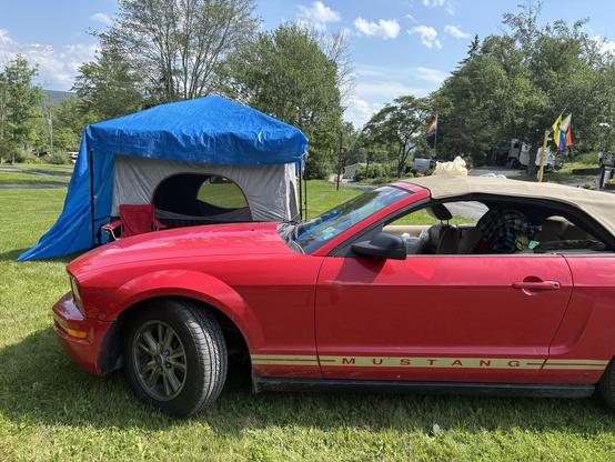 Side view, red mustang parked at a campsite. Large square tent with blue tarp on top. Red folding chair, blue sky, wispy clouds.