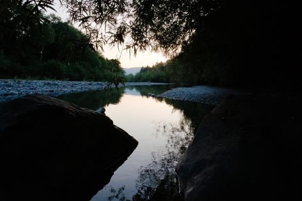 Foto eines Flusses, sitzend unter einem Baum aufgenommen. Die Äste des Baumes hängen am oberen Drittel des Fotos ins Bild, genau dort wo V-förmig der Horizont zu sehen ist. Der Fluss macht eine leichte S-Kurve, Links und rechts ganz vorne im Bild sind zwei große Felsen zu sehen. Weiter hinten links und rechts das Flussbett aus steinen, an beiden Seiten des Flussbetts stehen dicht an dicht Bäume.