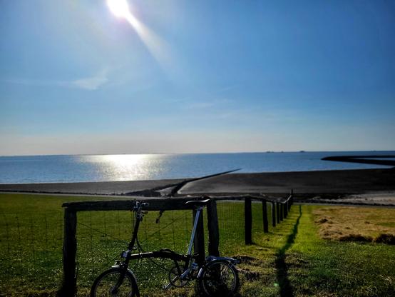Blick über Deichwiesen und Watt aufs weite Meer. Die im Bild oben links spiegelt sich die Sonne am fast wolkenlosen Himmel im blauen Meer. Im Bild oben rechts sind dunkle Punkte erkennbar von Land und Haus am entfernten Horizont. Im Bildvordergrund ist ein Brompton Fahrrad am Weidenzaun angelehnt.