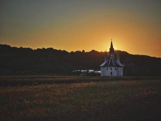A landscape-oriented photo shows a stone church with a bell tower and two smaller spires on the right side of the frame, silhouetted against an orange and yellow sunset. The sun is visible behind the church, close to the horizon line. In the foreground, a field of tall, dry grass stretches across the bottom of the image, with a darker, forested hill behind the church filling the rest of the background.
