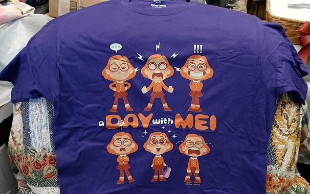A purple t-shirt with depictions of Meilin Lee from Turning Red showing six different emotions. Smugness, anger, and excitement on the top row, and annoyance, joy, and despair on the bottom row. In the middle of the two rows is the caption "A day with Mei".