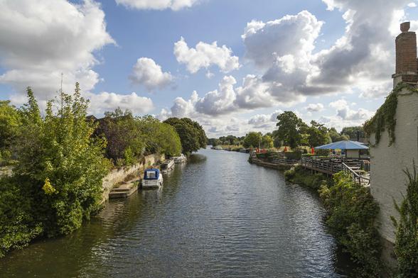 It's warm and sunny although the sky has plenty of billowing puffy white clouds that promise shady respite on occasion. We're standing on a bridge over a river perhaps twenty metres wide. The left bank of the river is largely lined with overhanging trees just behind a wall and a flat path against which small boats, canal boats, and narrow boats are moored. On the right bank there's the back of a pub with a large terrace on which there are benches, tables, chairs, and parasols. Further back are trees and fields and there are more boats of varying sizes moored along this bank too. It feels very peaceful.