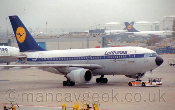 Side view of a twin engined jet airliner being towed from left to right by a low-slung white tug with a yellow stripe along the body, which has clasped and raised the plane's nosewheel in order to move it around.
The plane has a white upper body, with a grey belly, and a dark blue stripe running covering the passenger cabin windows.
There are dark blue "Lufthansa" titles on the upper forward fuselage, with the dark blue outline of a circle containing a stylised blue bird under the cockpit windows.
The white registration "D-AICB" is on the blue stripe at the rear of the fuselage, with black text "Airbus A310" on the lower rear body.
The tail is dark blue, with a yellow circle containing a stylised blue flying bird, similar to the logo under the cockpit.
Grey concrete apron fills the foreground, with grey buildings and white planes with blue and yellow tails filling the background under grey sky.