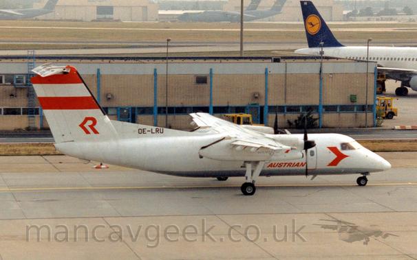 Side view of a high-winged, twin propellor-engined airliner taxiing from left to right.
The plane is mostly white, with a grey belly, and red "Austrian" titles on the lower forward fuselage, with a red arrowhead just below and aft of the cockpit.
There are a pair of thick red stripes at the top of the white tail, with a stylised red upper-case letter "R" at the base.
A pale brown building stretches across the background, with a white jet airliner with a blue and yellow tail just taxiing out of frame at the top right.
Large, green, 4 engined military cargo planes are clustered around a cream coloured hangar in the distance, slowly vanishing in to haze.