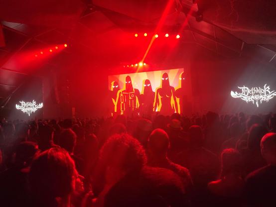 Dethklok in de Metal Dome op Graspop 2025. De band zie je eigenlijk niet, maar achter hen 5 donkere figuren met witte ogen op een lichtrode achtergrond. Er branden rode podiumlampen, en op de voorgrond zie je publiek.