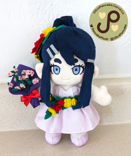 An 8 inch tall plush of Sayaka Maizono wearing her formal pink dress from her 10th Anniversary artwork. She has long, navy blue hair that is pulled into a high ponytail. She has decorative garlands of red and yellow flowers in her hair and around her waist. She's holding a bouquet of pink cherry blossoms.