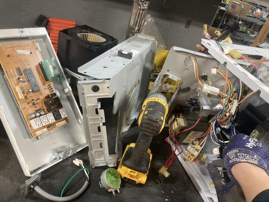 A mostly disassembled microwave, with my right hand holding it so it won’t fall off the workbench. There are circuit boards, capacitors, switches, etc. to be salvaged.