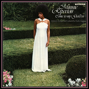 Minnie, en robe blanche dénudant ses bras, en afro, contemplant des fleurs roses et blanches dans un jardin à haies basses bien taillées