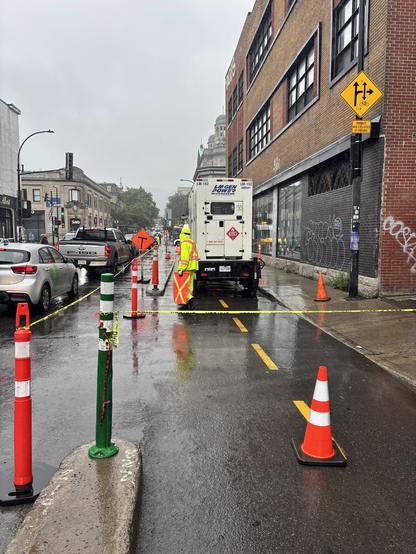 Scène de rue par temps pluvieux présentant une zone de construction. Des cônes de signalisation et du ruban adhésif bloquent une partie de la route où est garé un véhicule de service. Des ouvriers vêtus de vêtements de pluie brillants sont présents. Les bâtiments avoisinants présentent une architecture ancienne en briques et des éléments modernes.