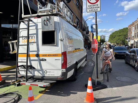 Une camionnette utilitaire blanche surmontée d'une échelle est garée à côté d'une piste cyclable. Un cycliste casqué et en tenue décontractée est arrêté près d'un panneau stop. La rue est bordée d'arbres et de bâtiments, et d'autres véhicules sont visibles à l'arrière-plan.