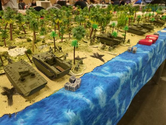 a close up of some miniature landing craft, troops, and dice used in the game over a blueish table cloth to represent the water next to the beach. on the right side are two tape measures.