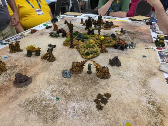 a 1.5m by 1.5m tan/desert type post apocalyptic terrain setup for Gaslands Capture the Flag. Most of the carts shown are tiny monster trucks with large tires, and a Warhammer 40k standard is used as each team's flag.