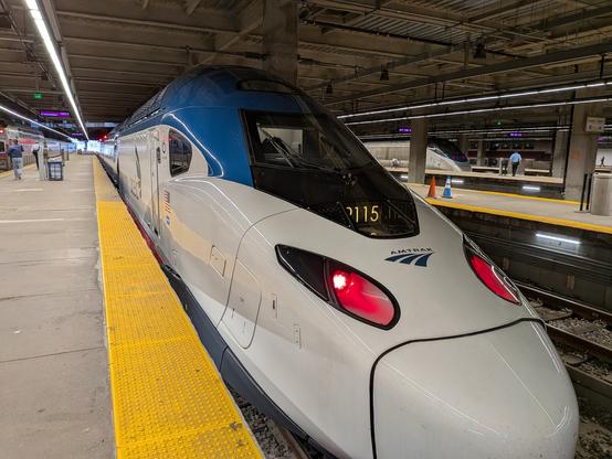 The front of a high speed train, with a sleek aerodynamic nose cone leading to a long set of rail cars. It is at station, and is styled in white and dark blue with red accents. Two tracks behind it is an older looking train of a comparable but more antiquated vintage.
