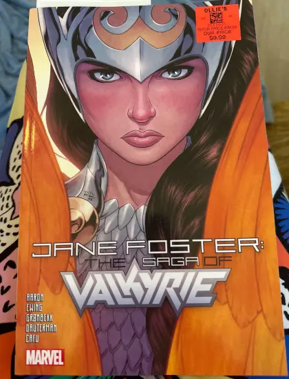 A comic book as seen from above. The title reads “Jane Foster: the Saga of Valkyrie.” The cover art depicts a close up of Jane facing forward. She’s wearing a silver and gold helmet. Her armor is the same colors. Two golden feathered wings are positioned in front of her slightly. Her hair is dark brown and her eyes are blue.